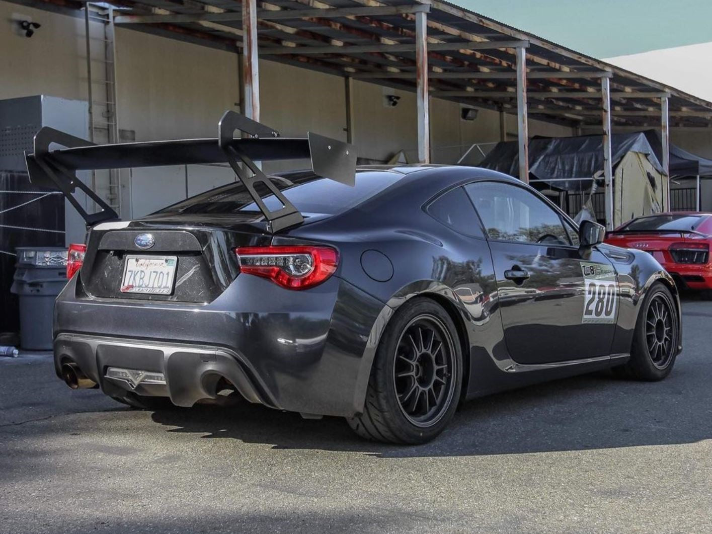 Rear three-quarter view of a dark gray FRS/BRZ motorsport car with GT wing near a warehouse