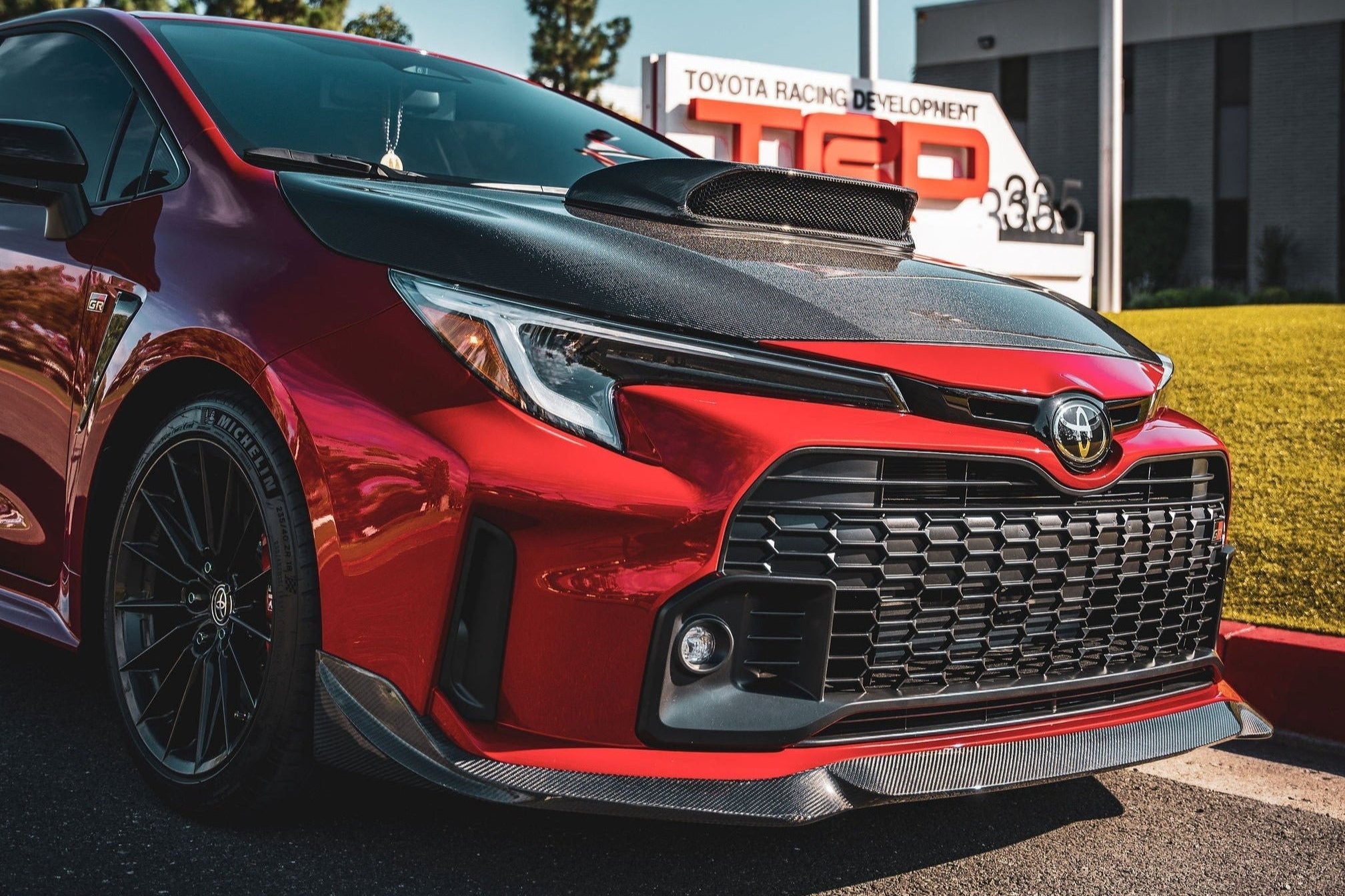 Front three-quarter view of a red GR Corolla with front lip parked outside a building