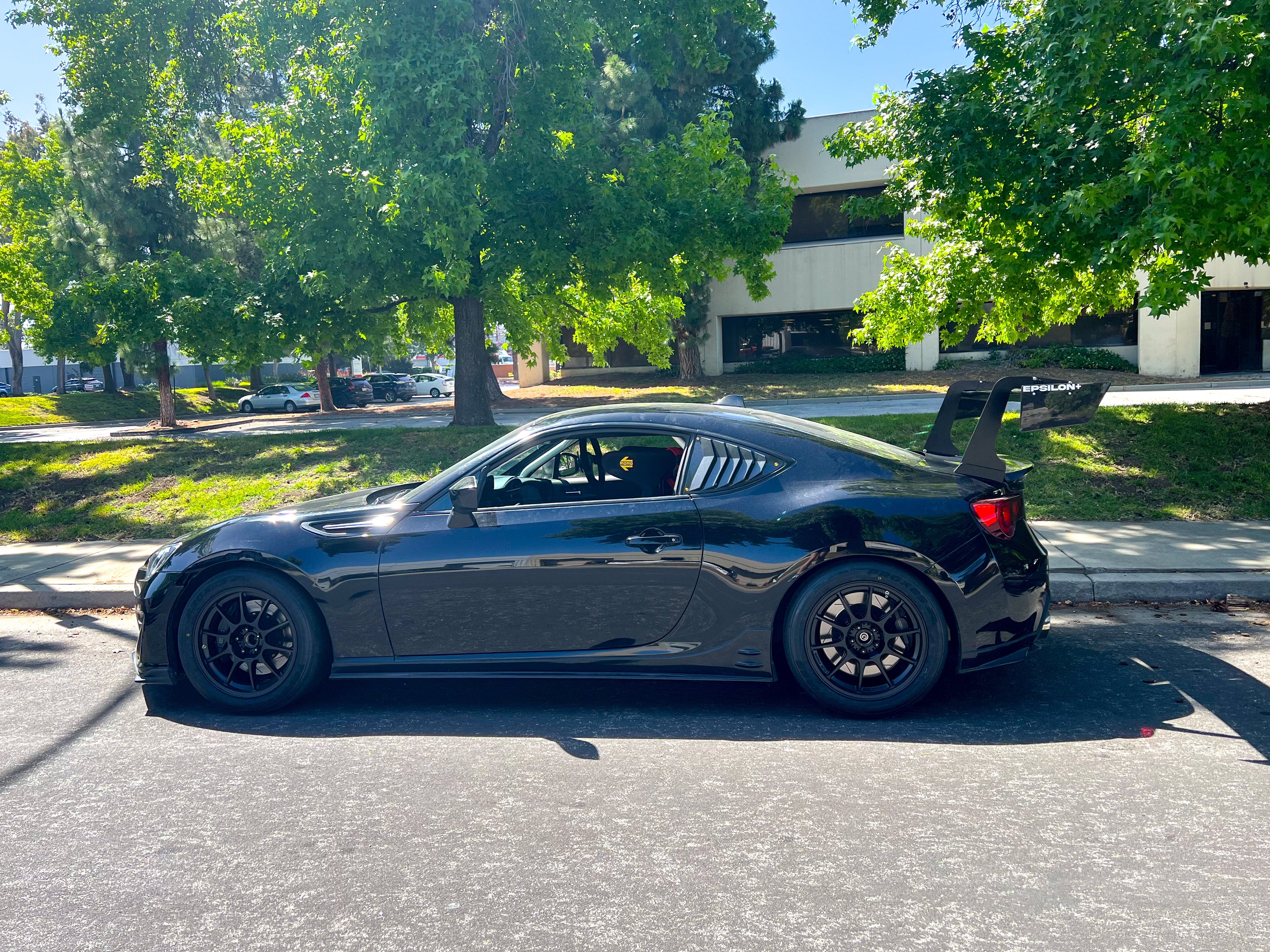 Front three-quarter view of a dark gray FRS/BRZ with GT wing on a tree-lined street