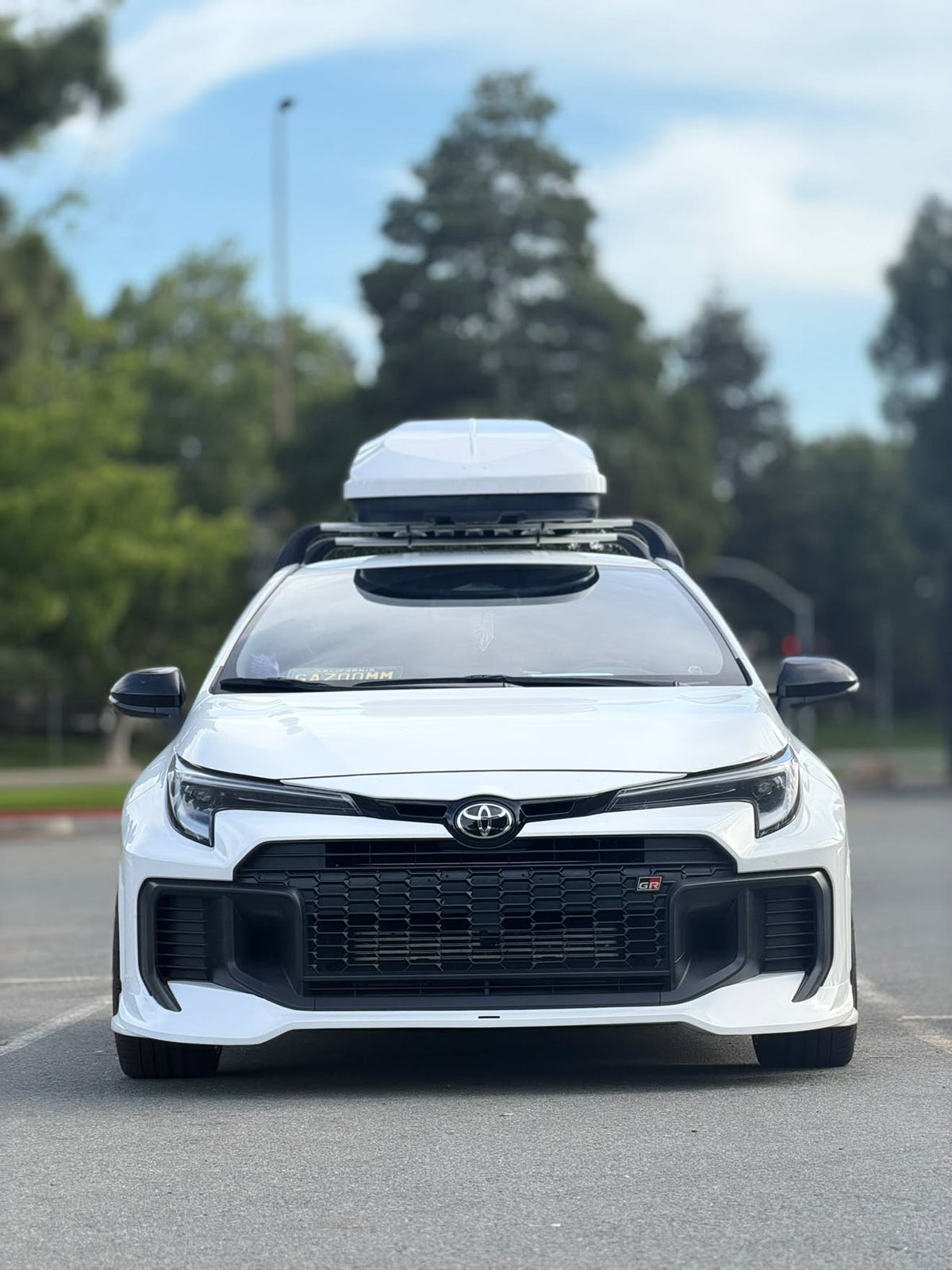 Front-on view of a white GR Corolla Kouki with front lip and roof box in a parking lot