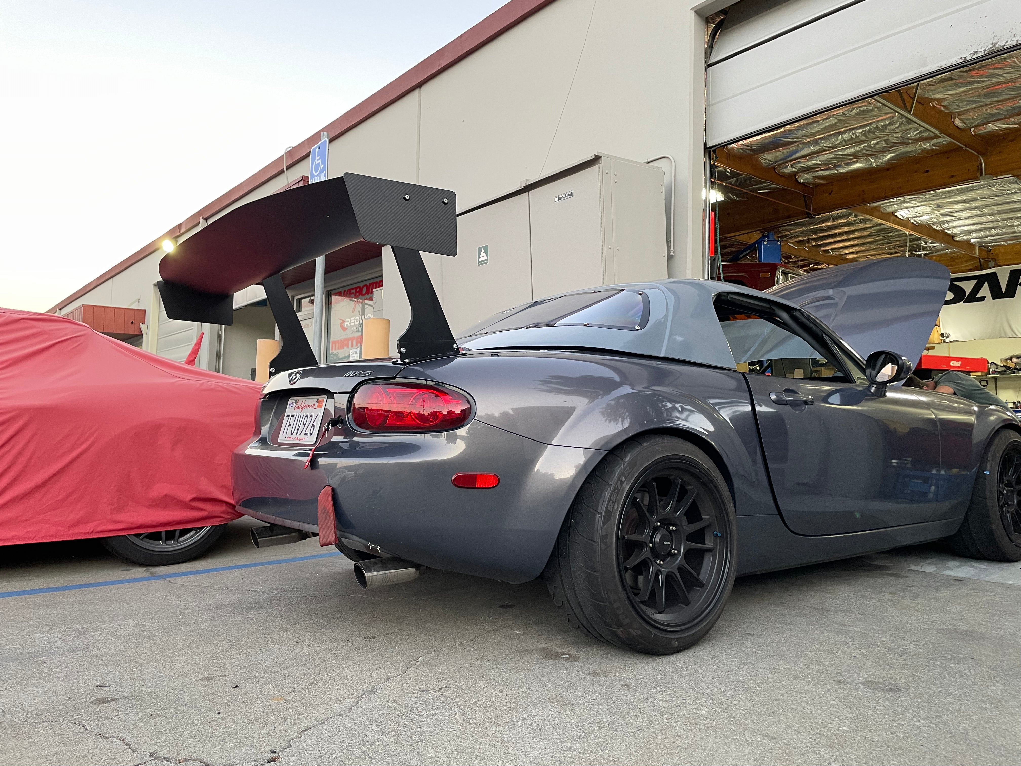 Rear three-quarter view of a gray Miata NC with GT wing beside a warehouse