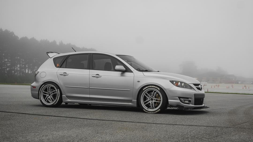 Side profile of a silver Mazdaspeed3 Gen1 with front splitter, foggy track background