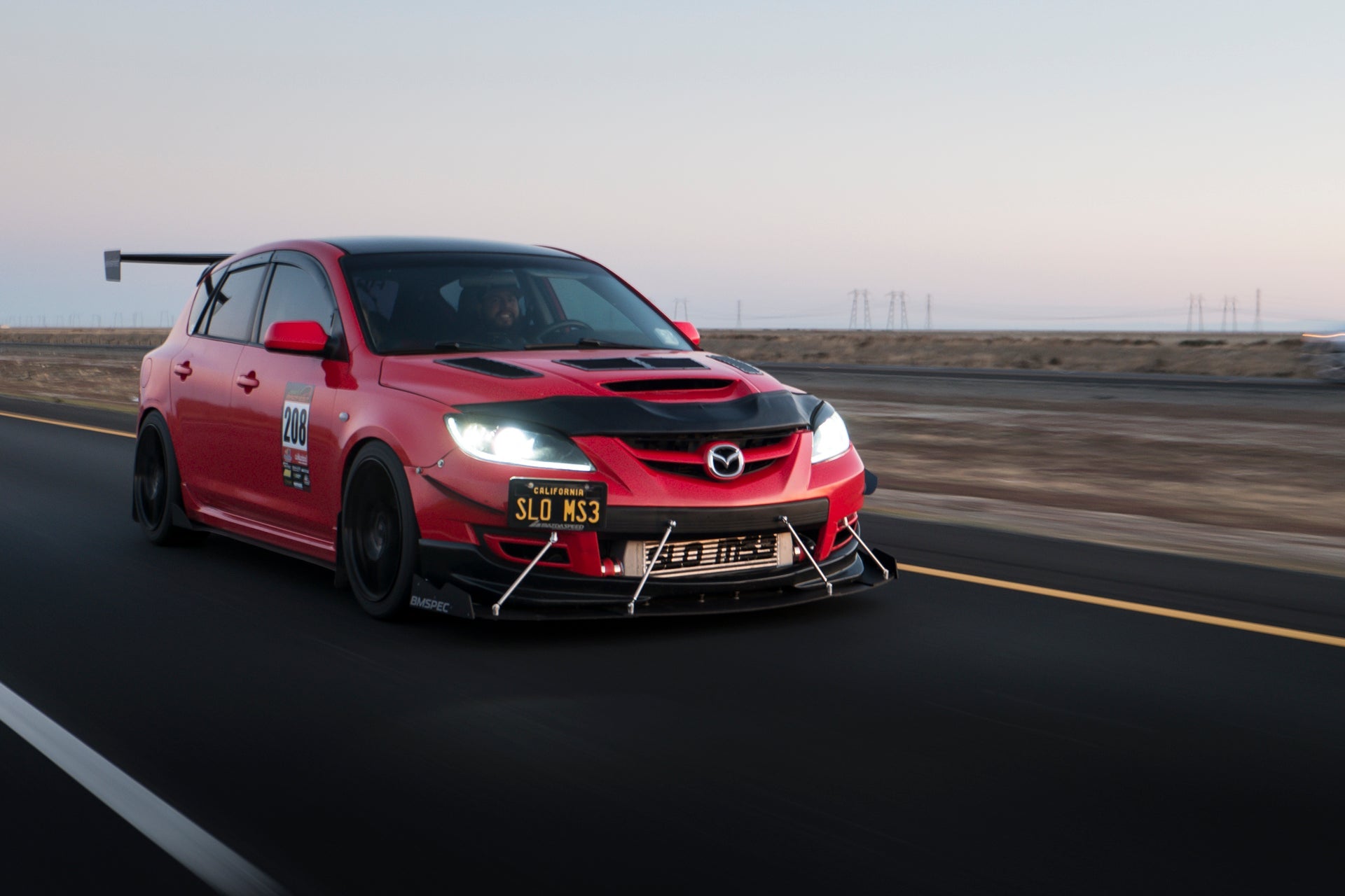 Front three-quarter view of a red Mazdaspeed3 Gen1 with GT wing at dusk on a road