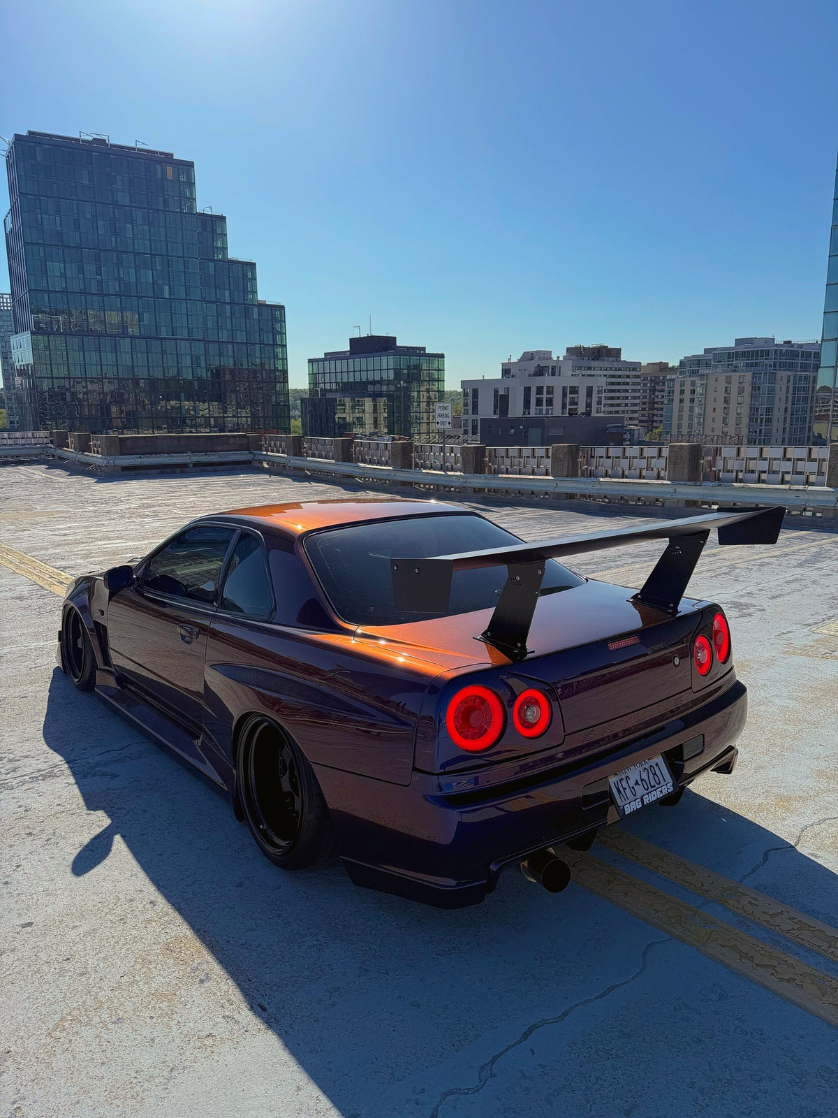 Rear three-quarter view of a bronze-to-purple Nissan Skyline R34 with an Epsilon+ GT wing on bottom-mount uprights, city skyline behind