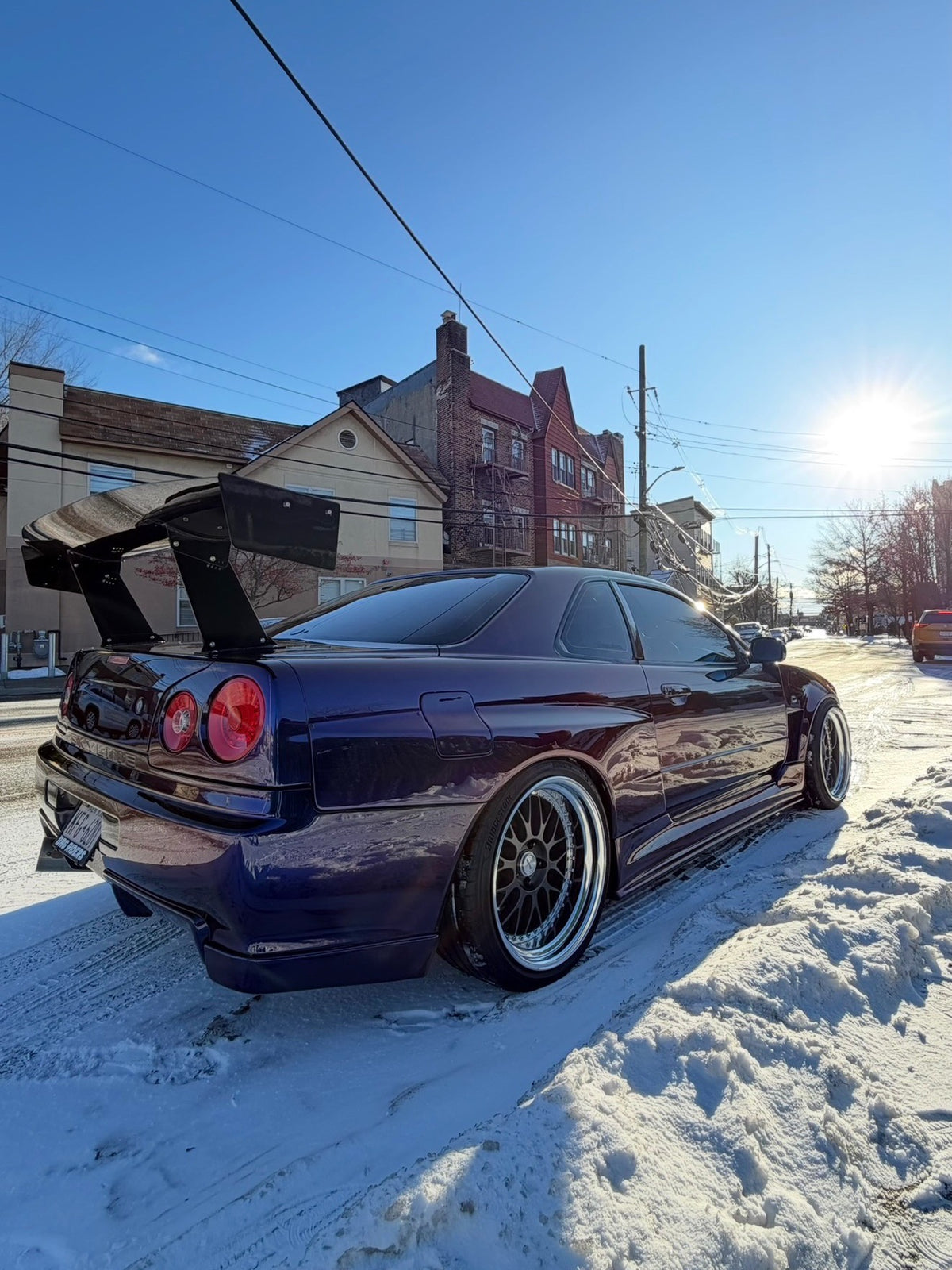 Low rear three-quarter view of a purple Nissan Skyline R34 with an Epsilon+ GT wing parked on a snowy street
