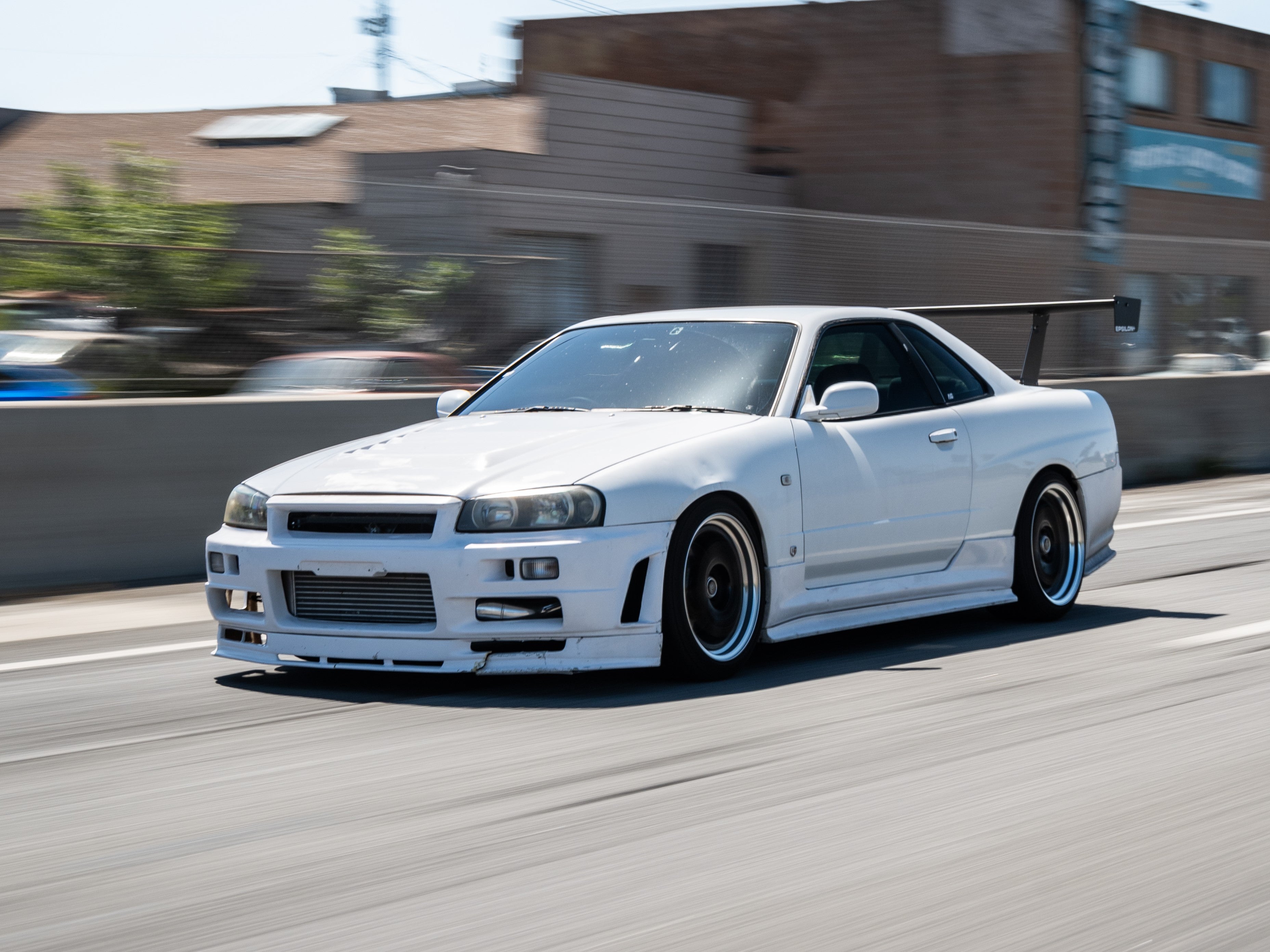 White Skyline R34 with GT wing driving past an industrial building