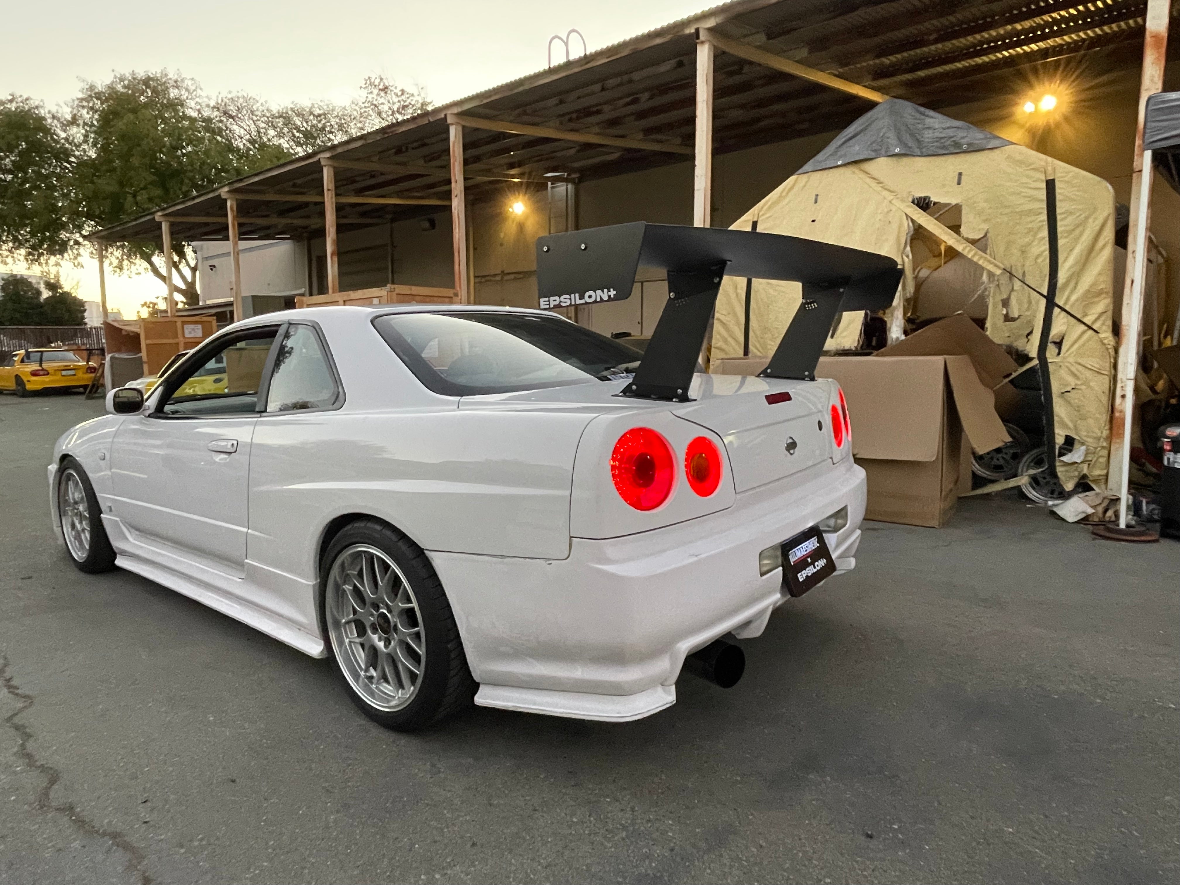 Rear view of a white Skyline R34 with the Epsilon+ GT wing under a carport