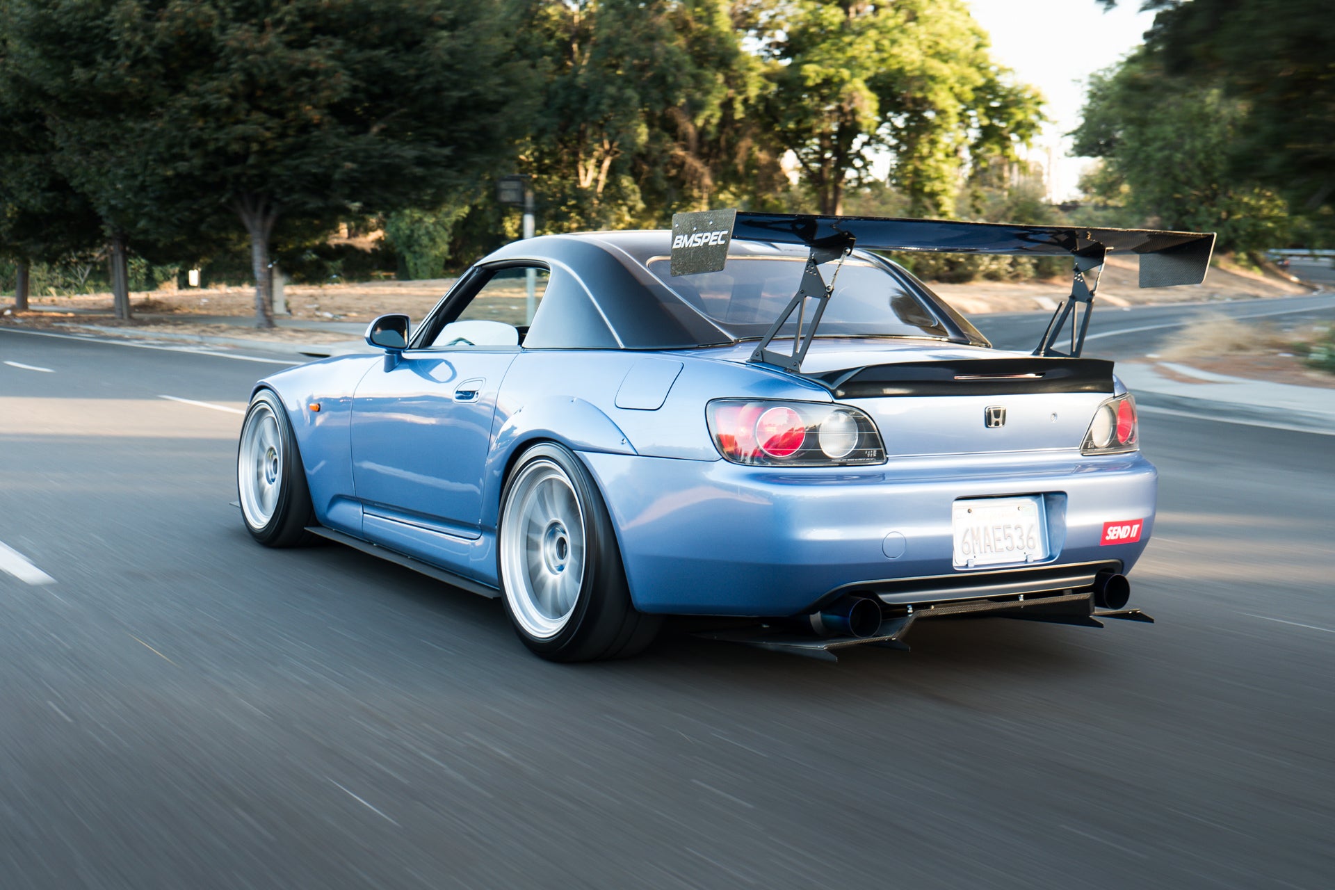 Blue Honda S2000 with V1+ rear diffuser and GT wing, rolling shot on a tree-lined road