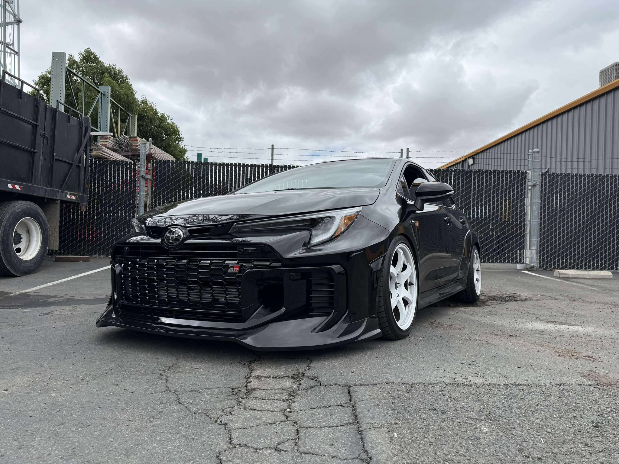 Front three-quarter view of a black GR Corolla Kouki with front lip near a warehouse