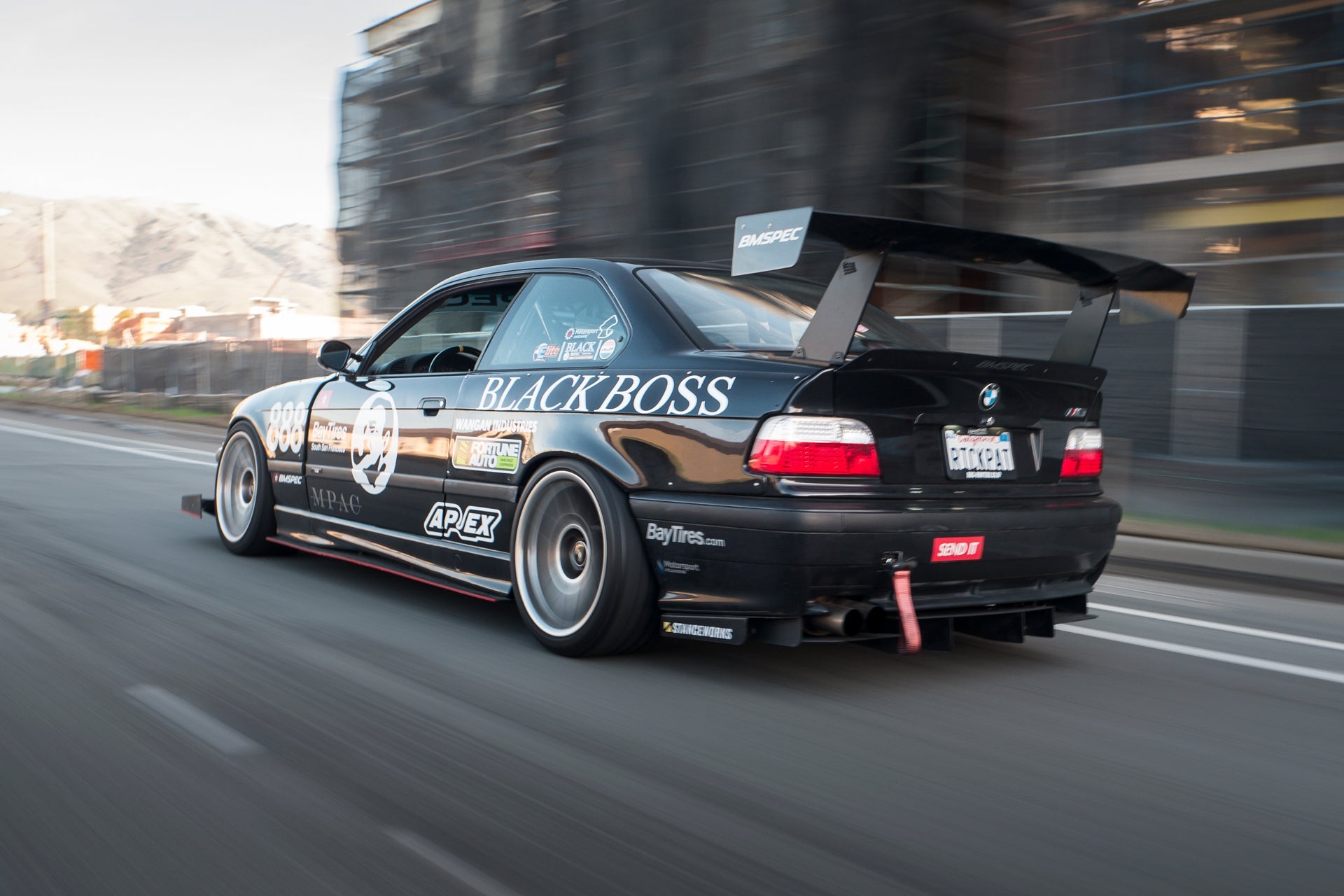 Black BMW E36 M3 motorsport car with GT wing driving through a city street at dusk