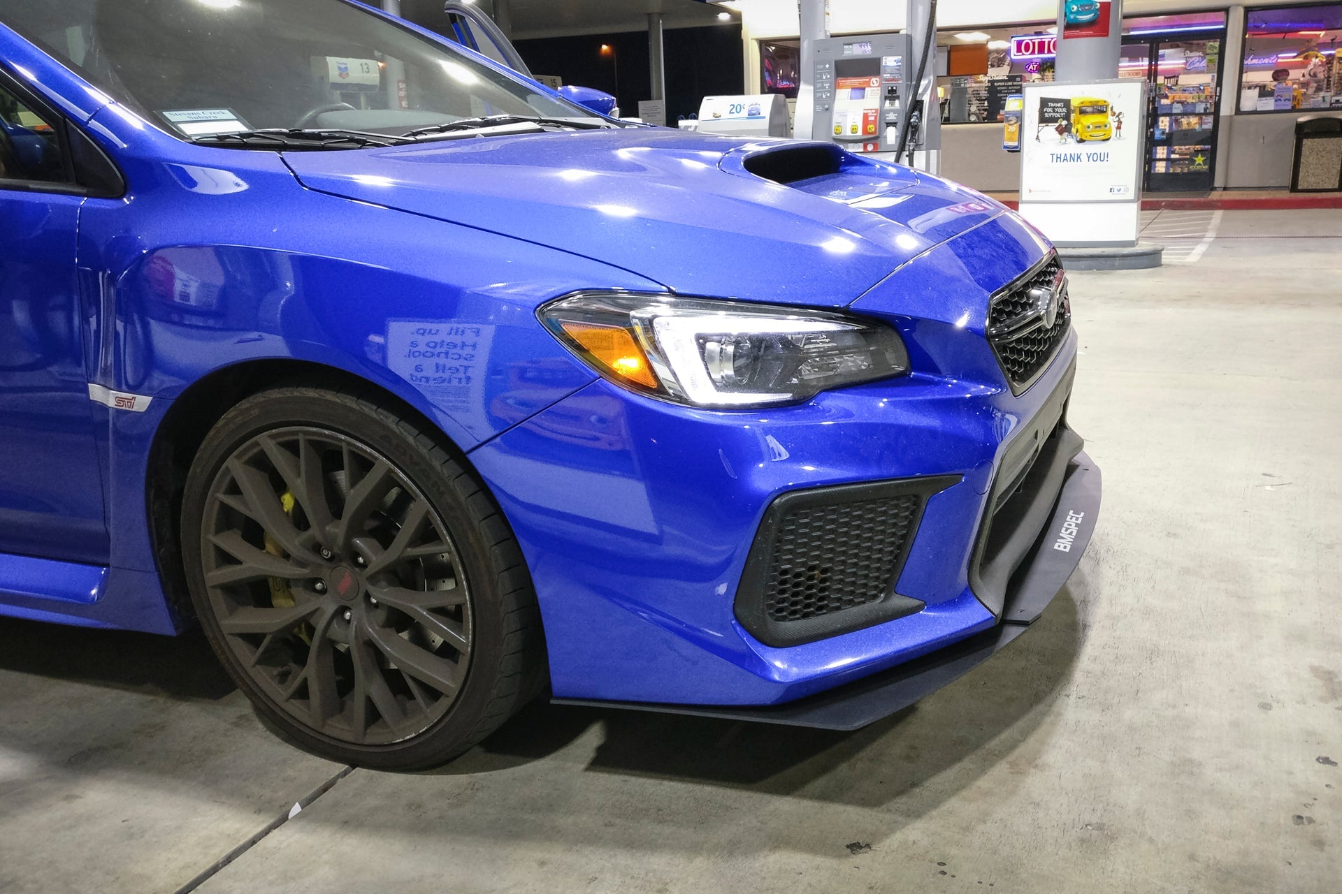 Close-up front fender view of a blue Subaru WRX STI with front lip at a gas station