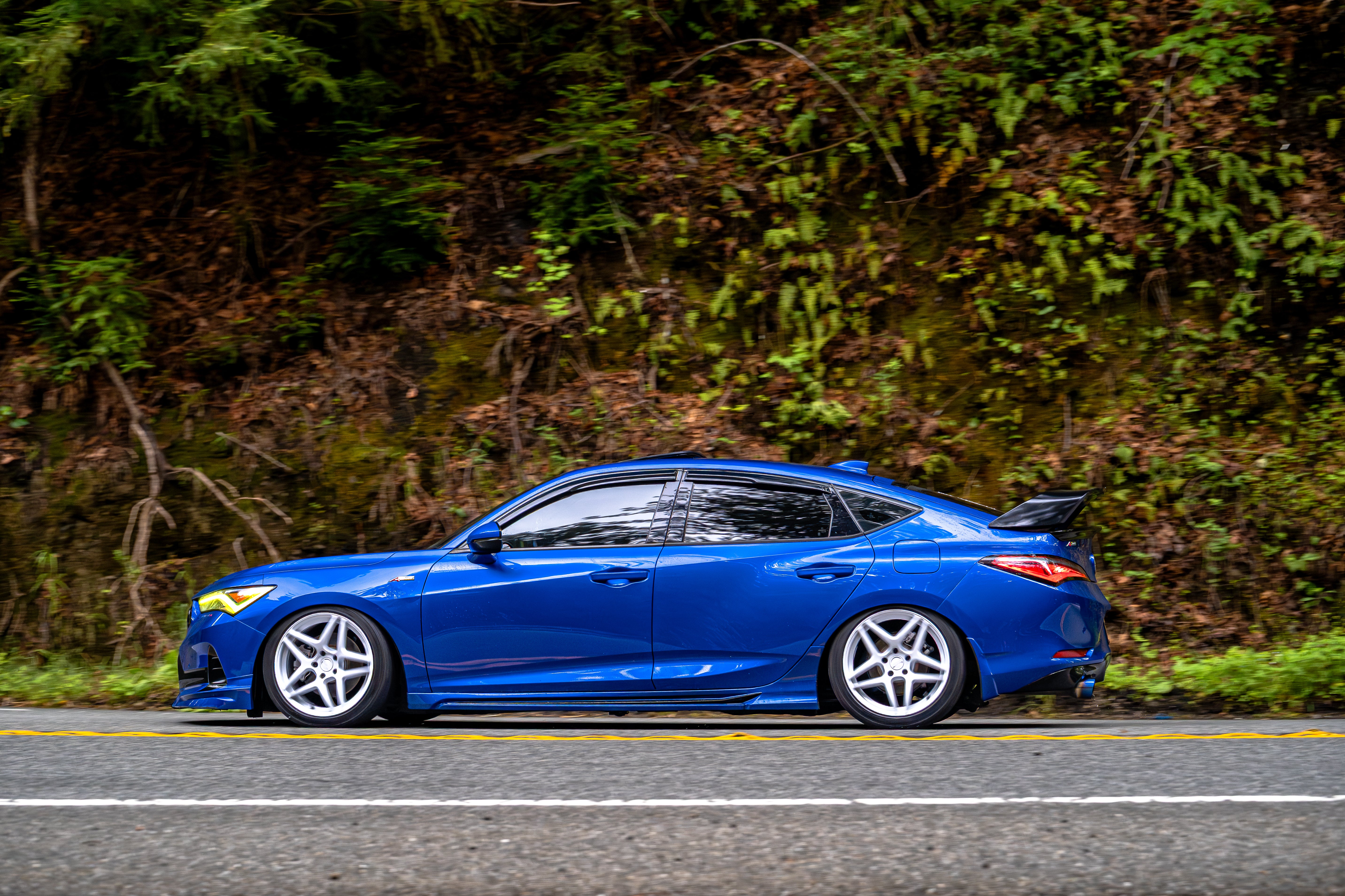 Side profile of a blue Acura Integra with the Sport Wing on a hillside road