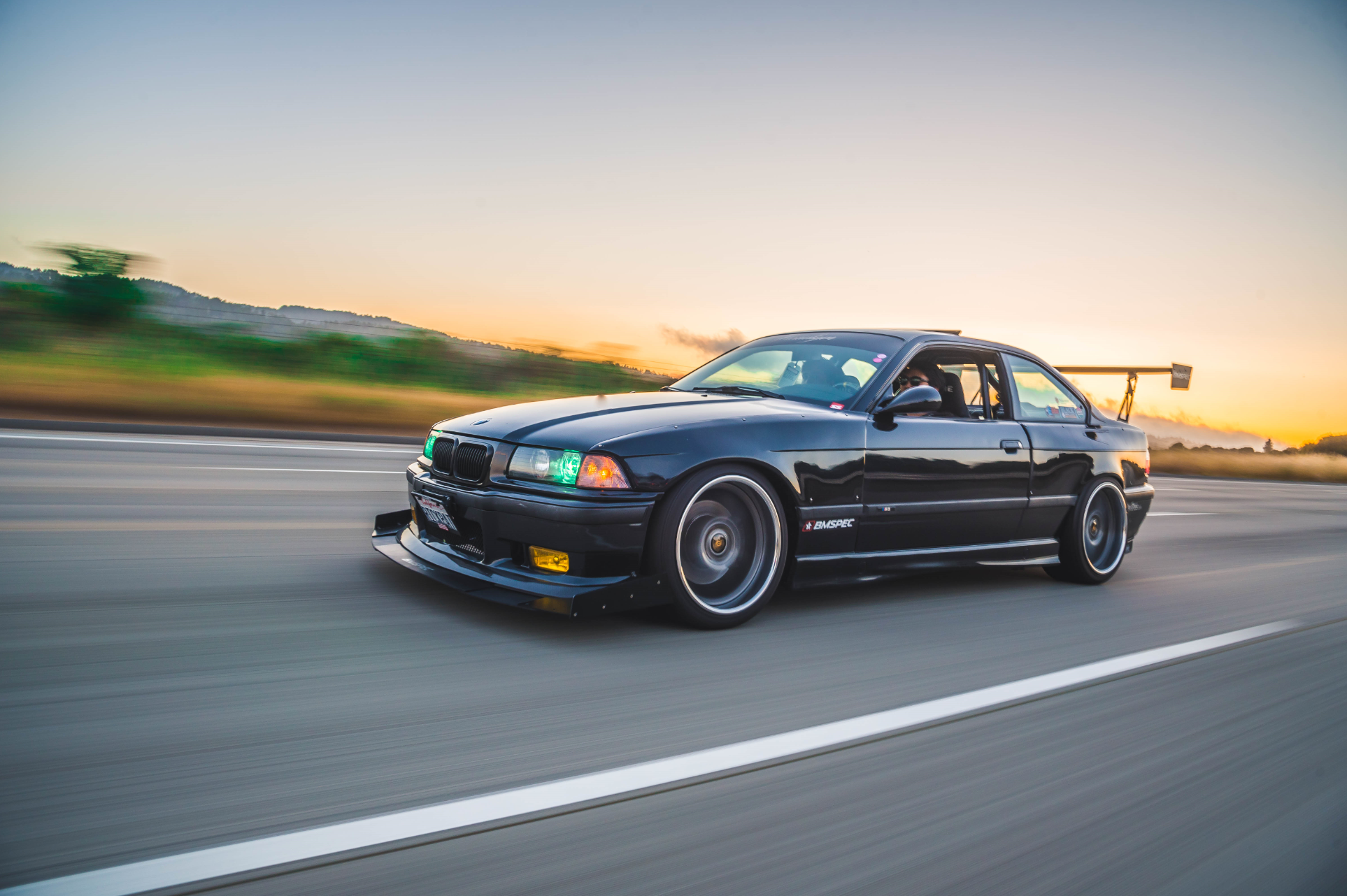 Black BMW E36 M3 with front splitter and GT wing driving on a highway at sunset
