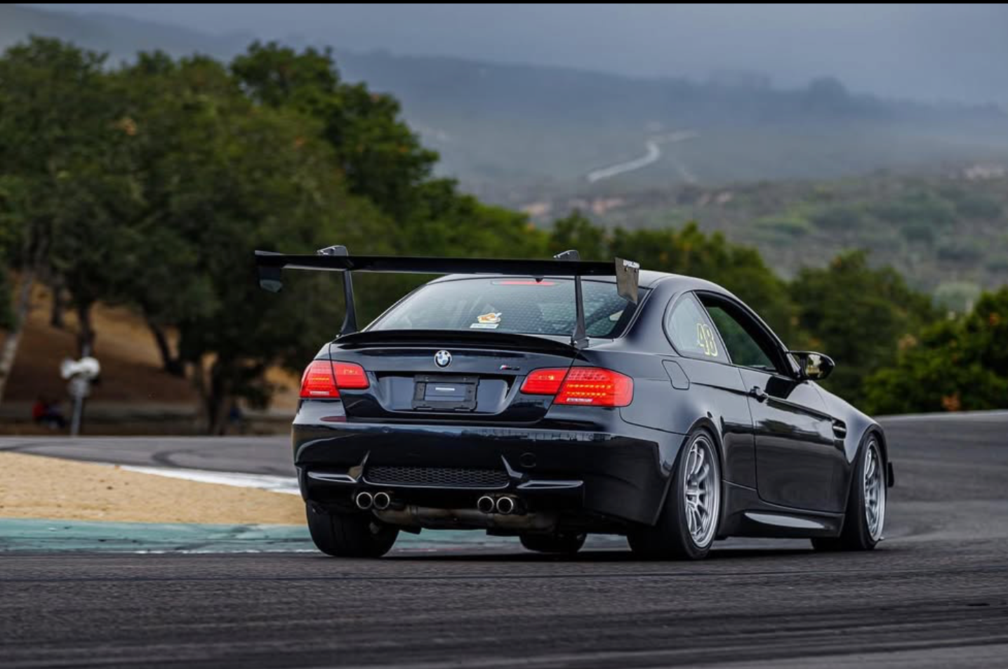 Black BMW E92 M3 with GT wing on a road course with hills behind