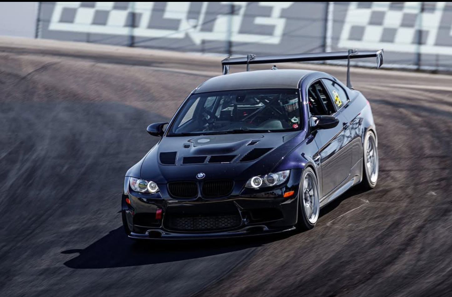 Black BMW E92 M3 with GT wing cornering on track, front three-quarter view