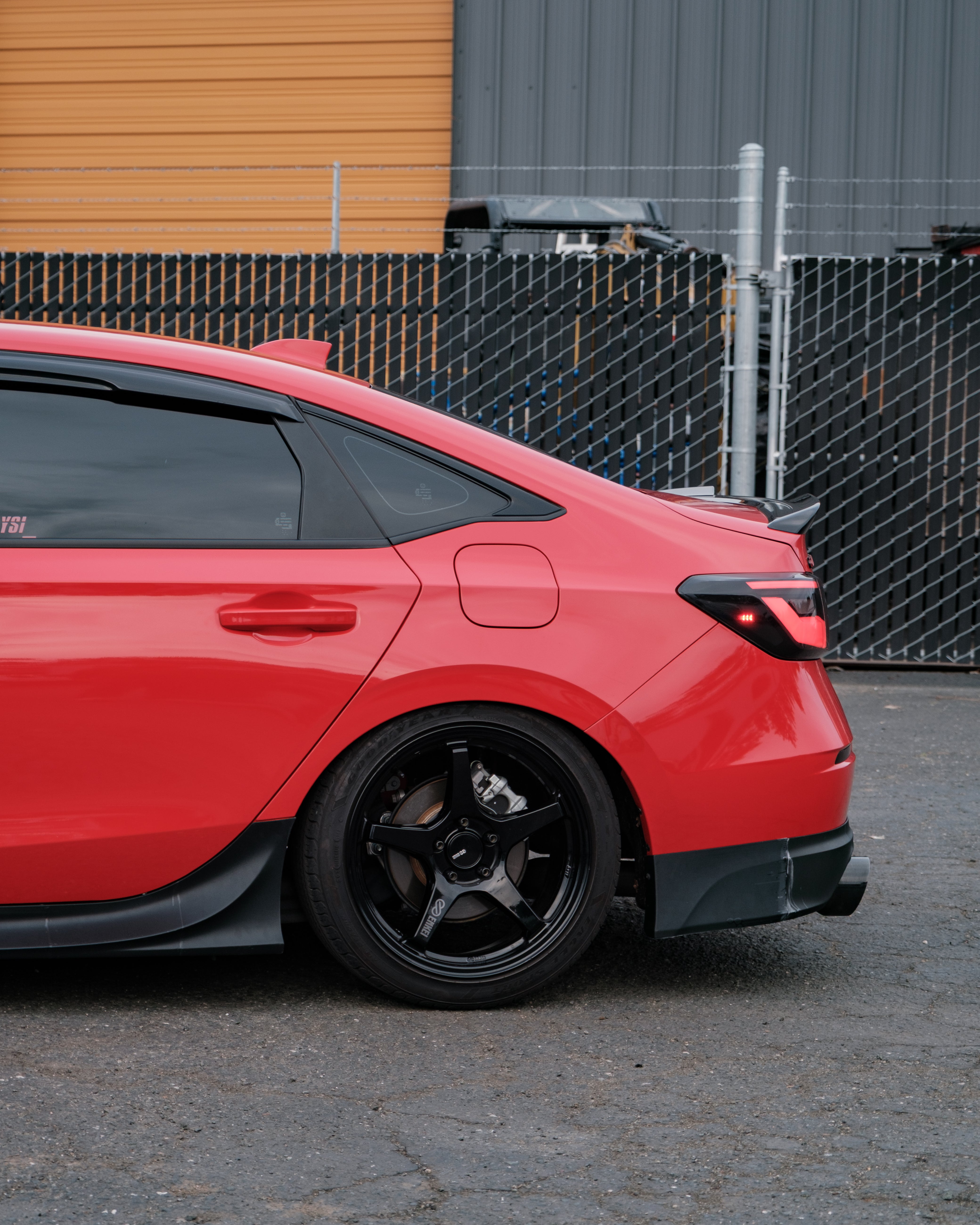 Side-rear view of a red Honda Civic Sedan with rear lip beside a fence