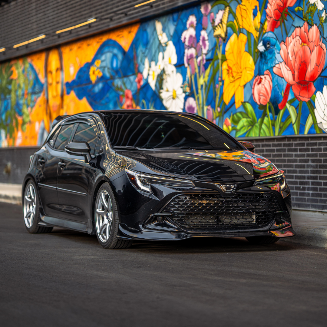 Black Toyota Corolla Hatchback Kouki with Epsilon+ front lip beside a floral mural