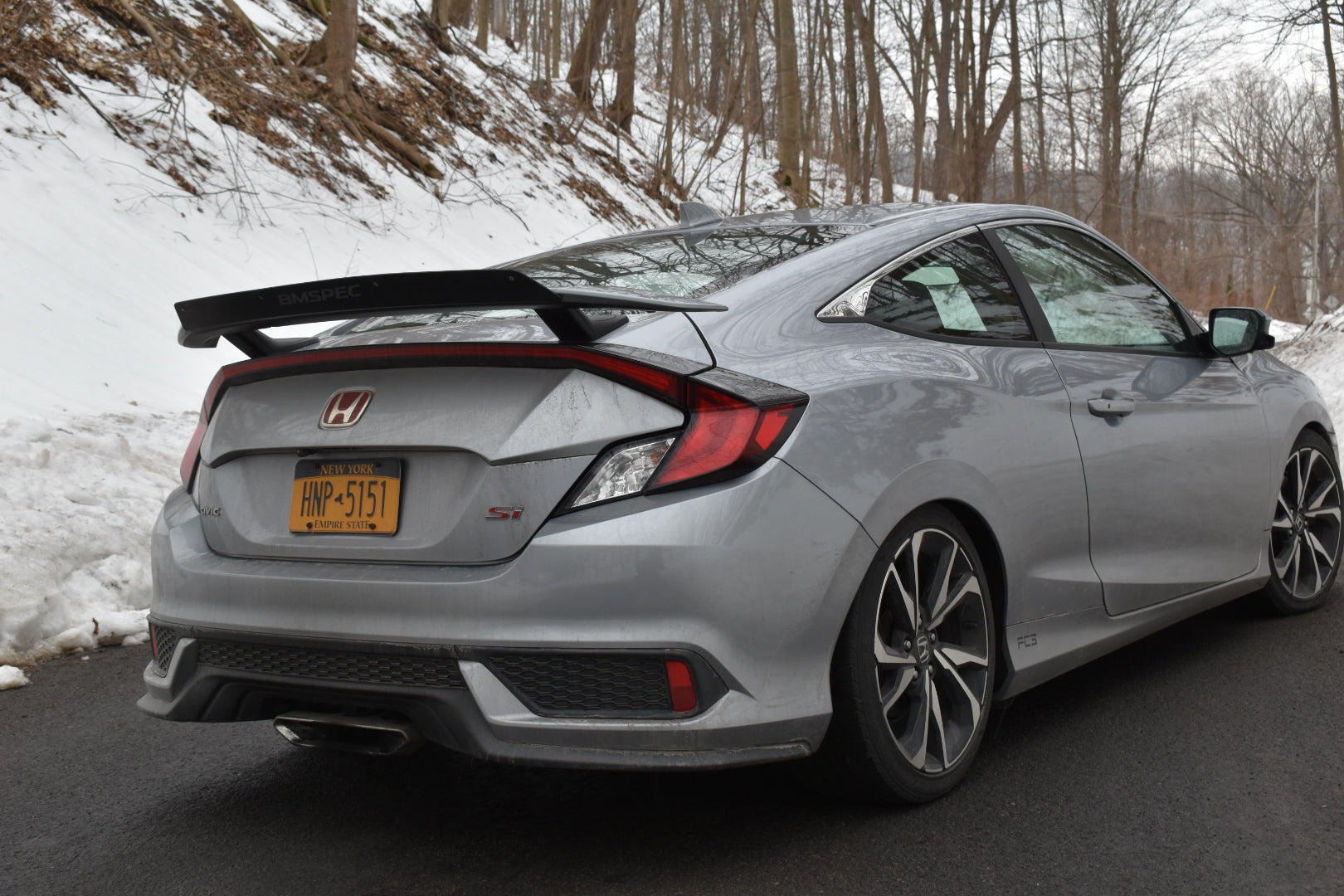 Rear three-quarter view of a silver Civic Si Coupe with Epsilon+ spoiler extension on a mountain road