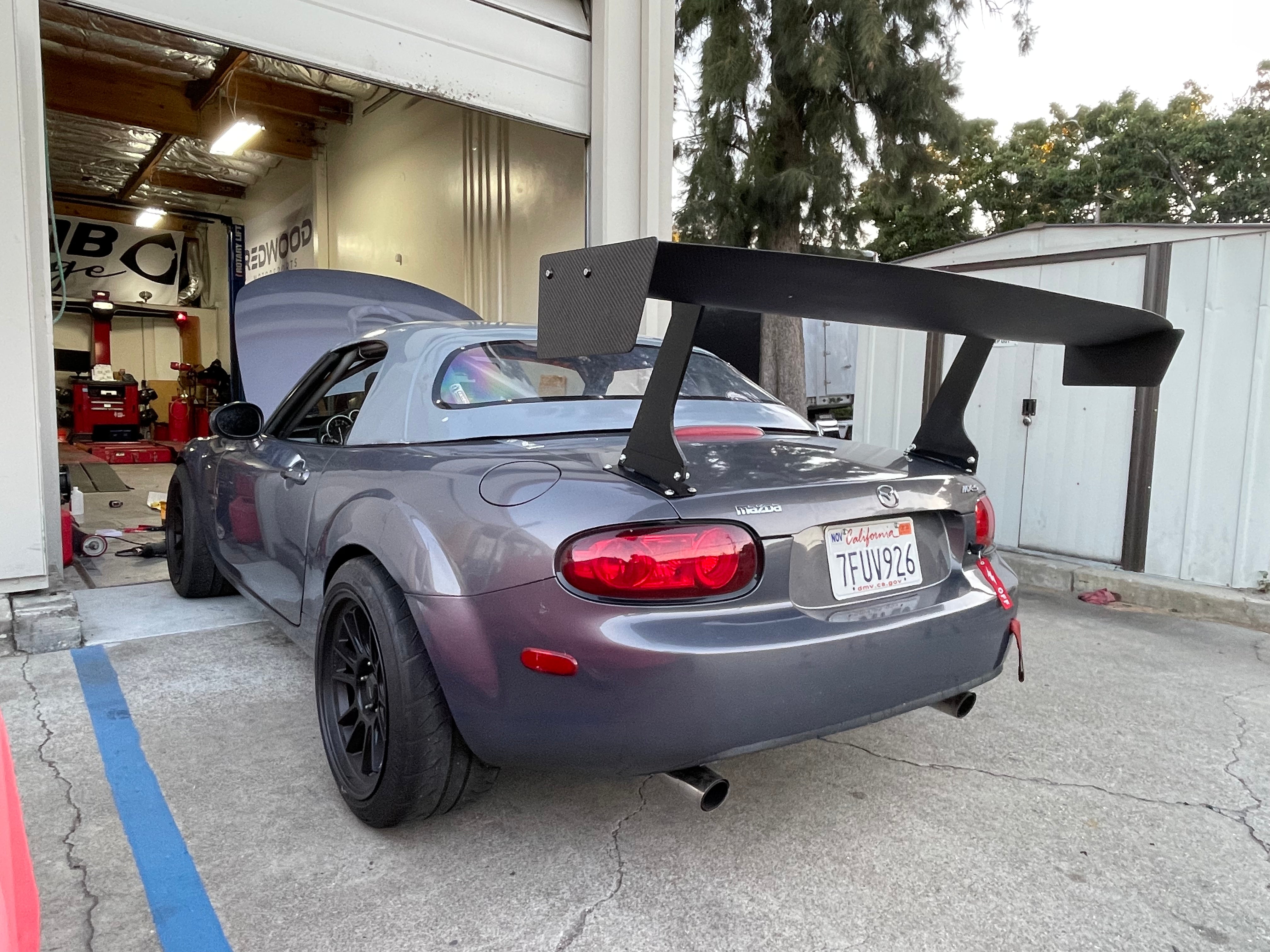 Rear three-quarter view of a gray Mazda MX-5 Miata NC with GT wing in the shop