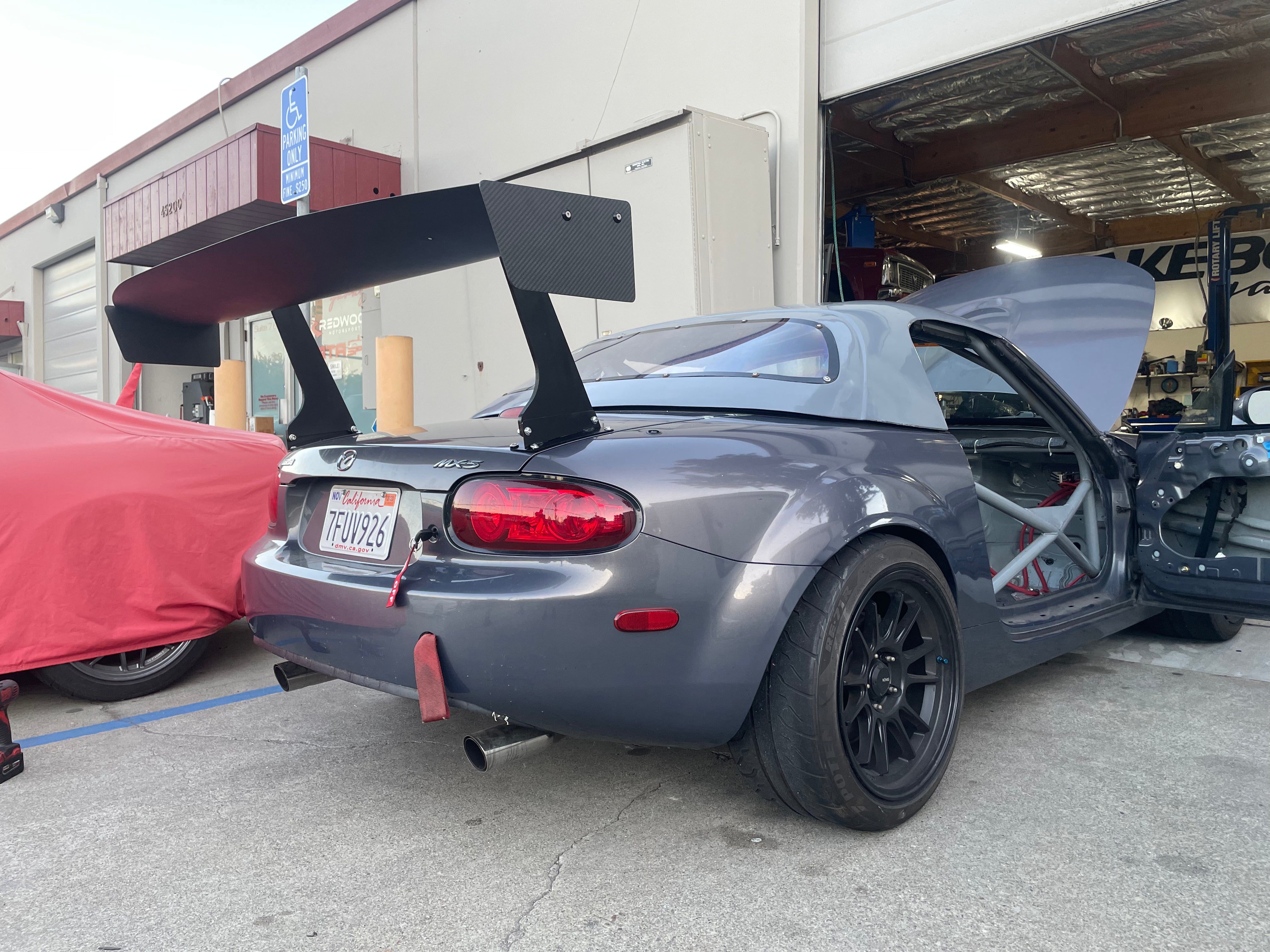 Rear three-quarter view of a gray Miata NC with GT wing parked outside