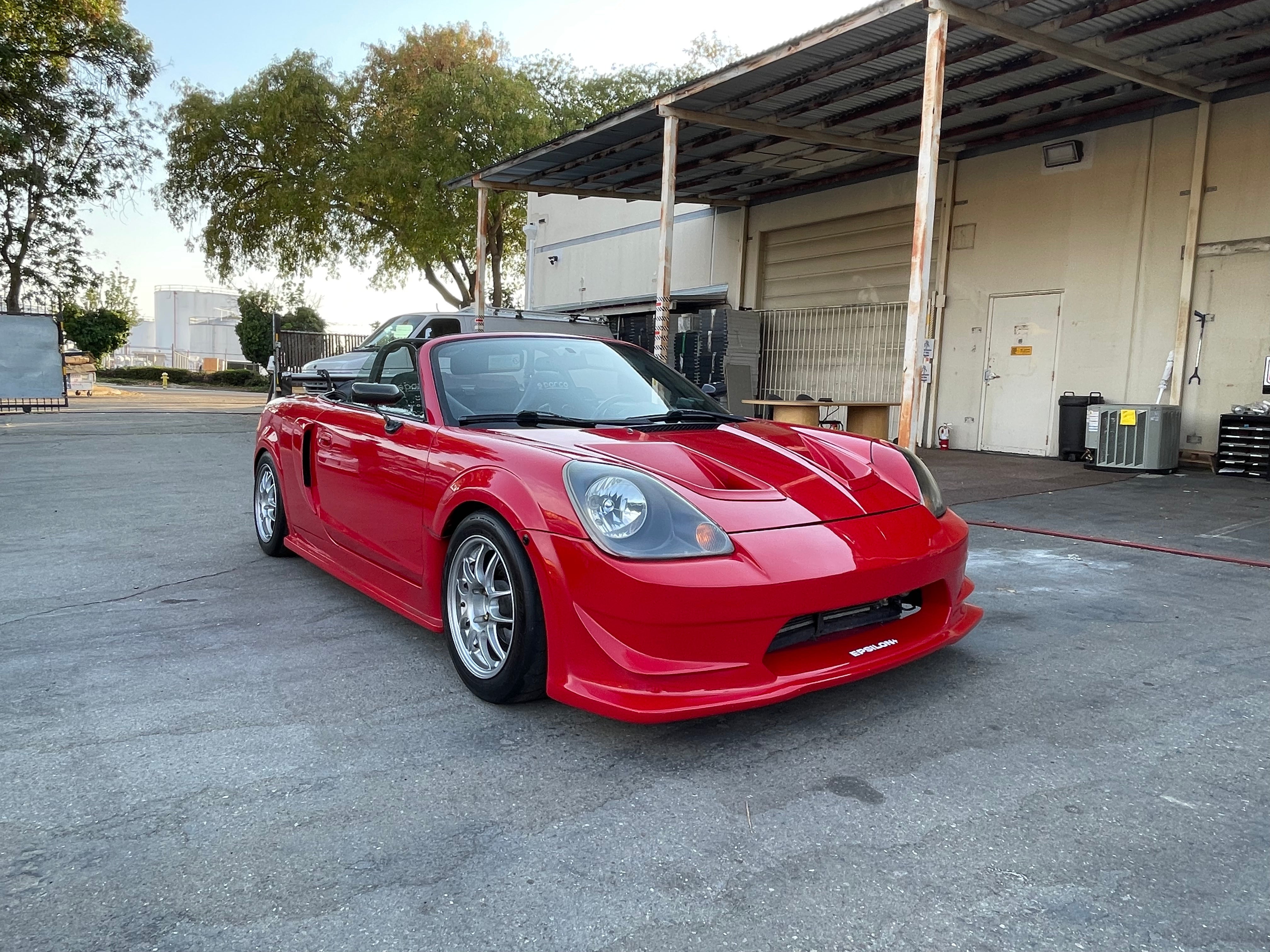 Front three-quarter view of a red MR2 Spyder front bumper beside a warehouse