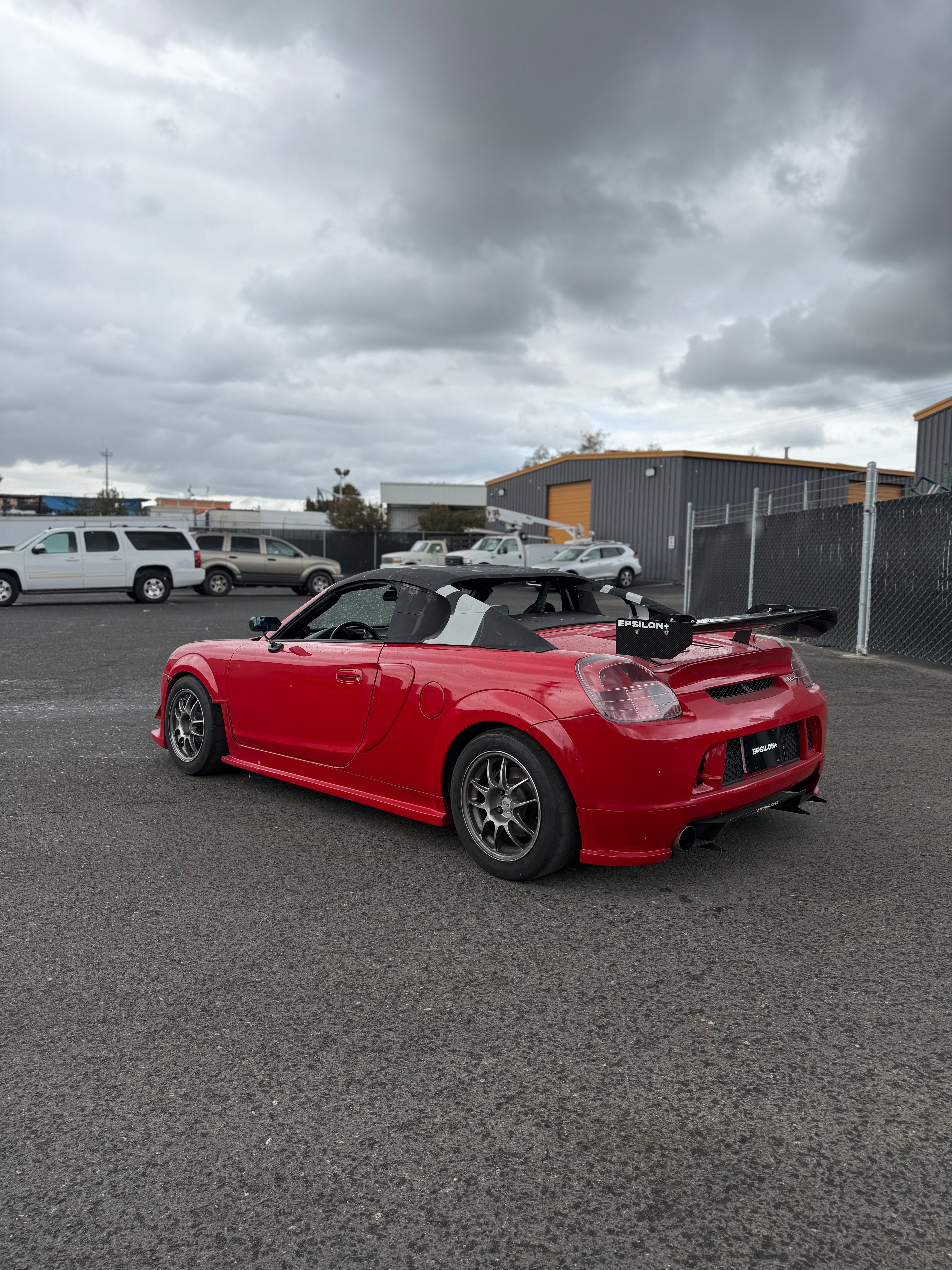Side profile of a red Toyota MR2 Spyder in a parking lot with hardtop hardware fitted