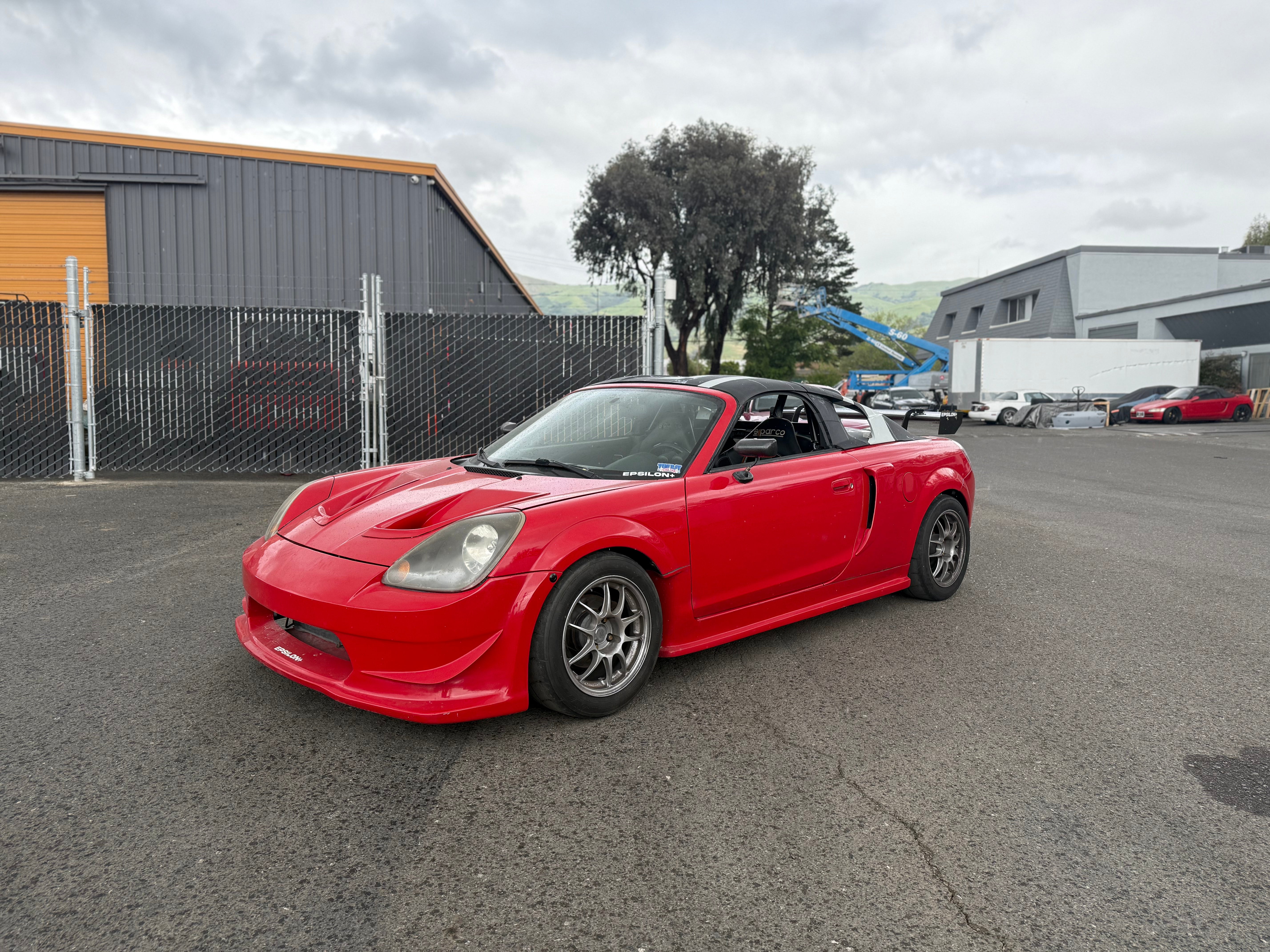 Front three-quarter view of a red Toyota MR2 Spyder parked by a warehouse