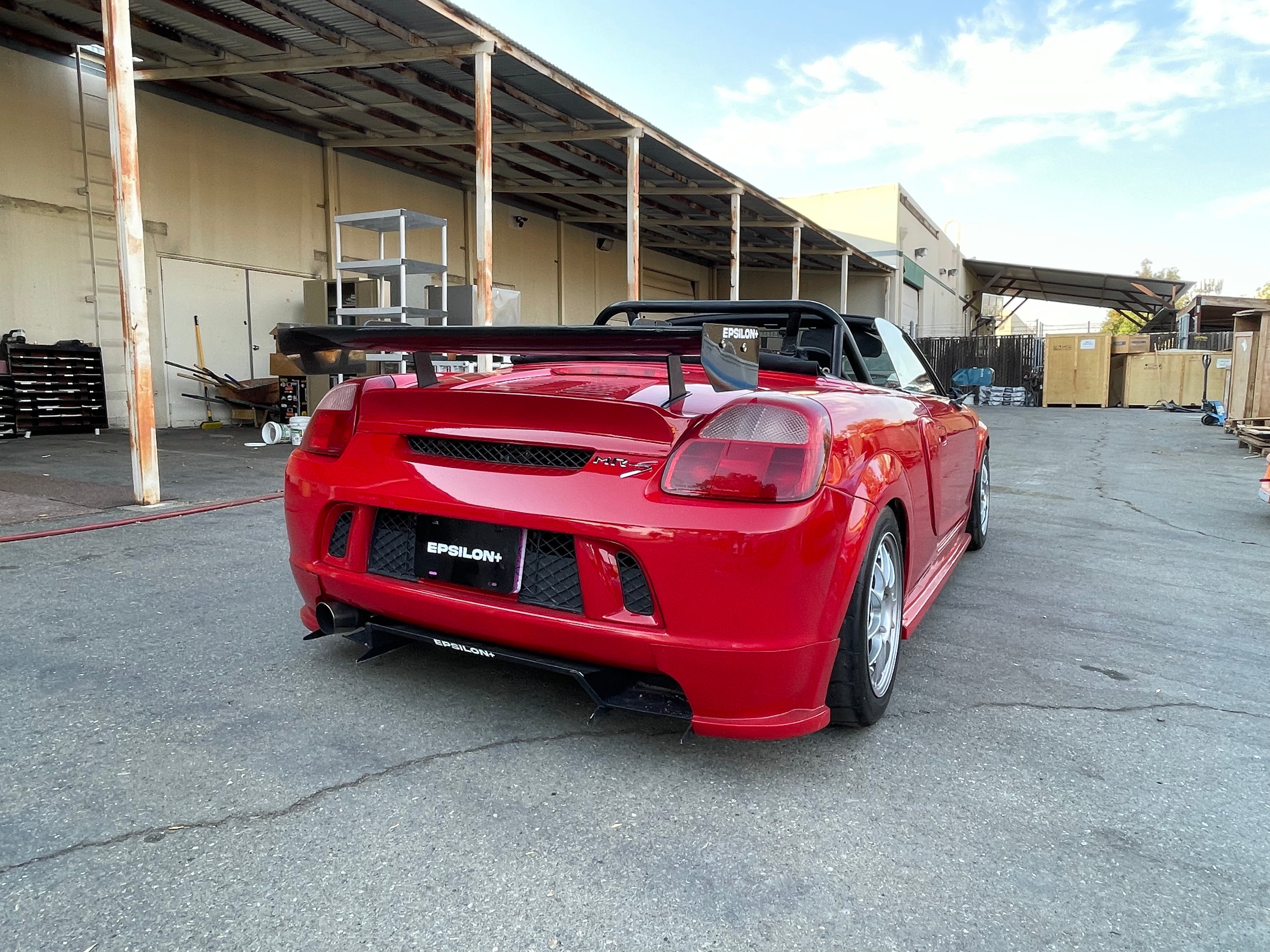 Rear three-quarter view of a red MR2 Spyder with rear bumper spats outside a warehouse