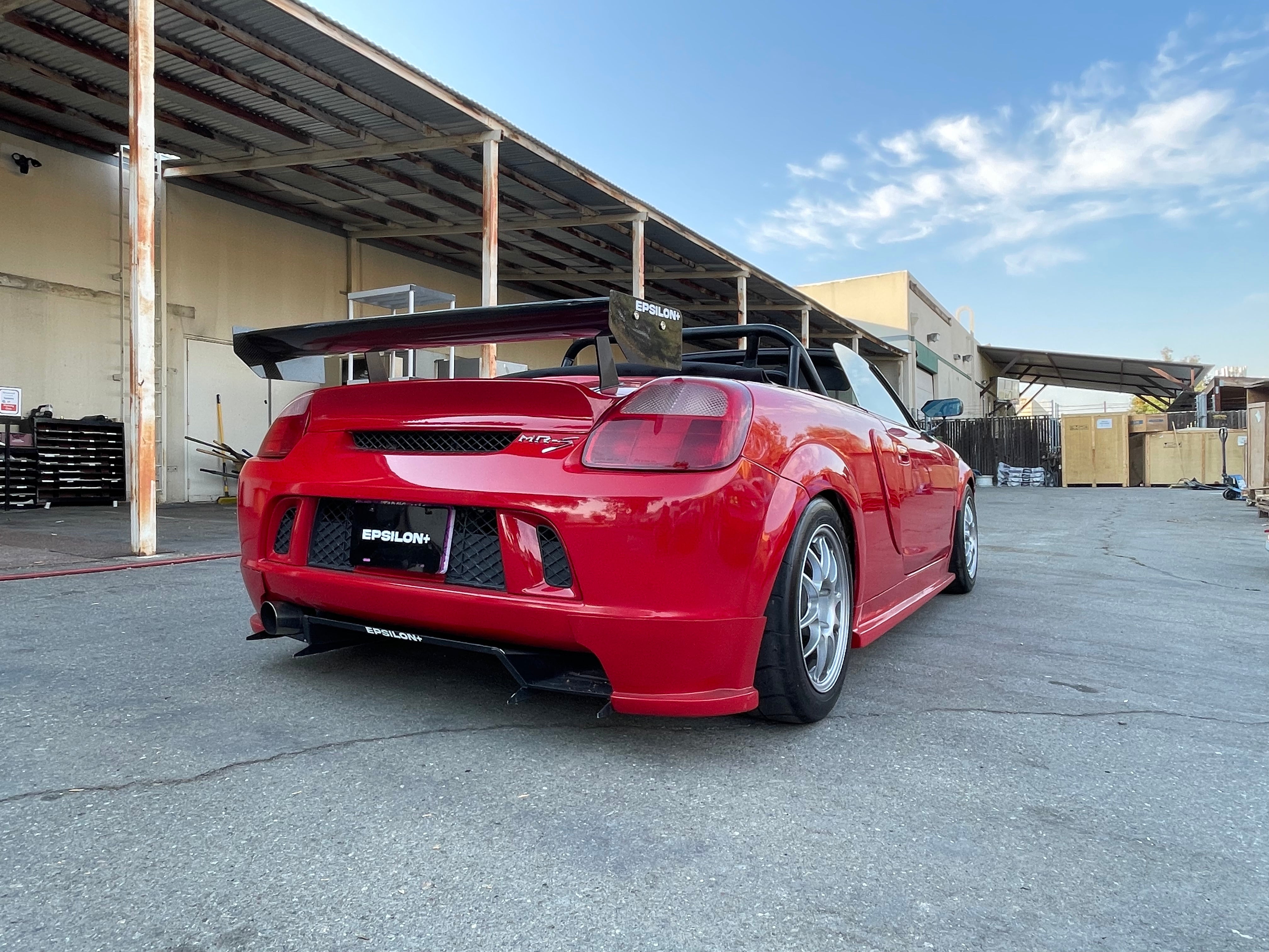 Rear three-quarter view of a red MR2 Spyder with rear wing parked outside a warehouse
