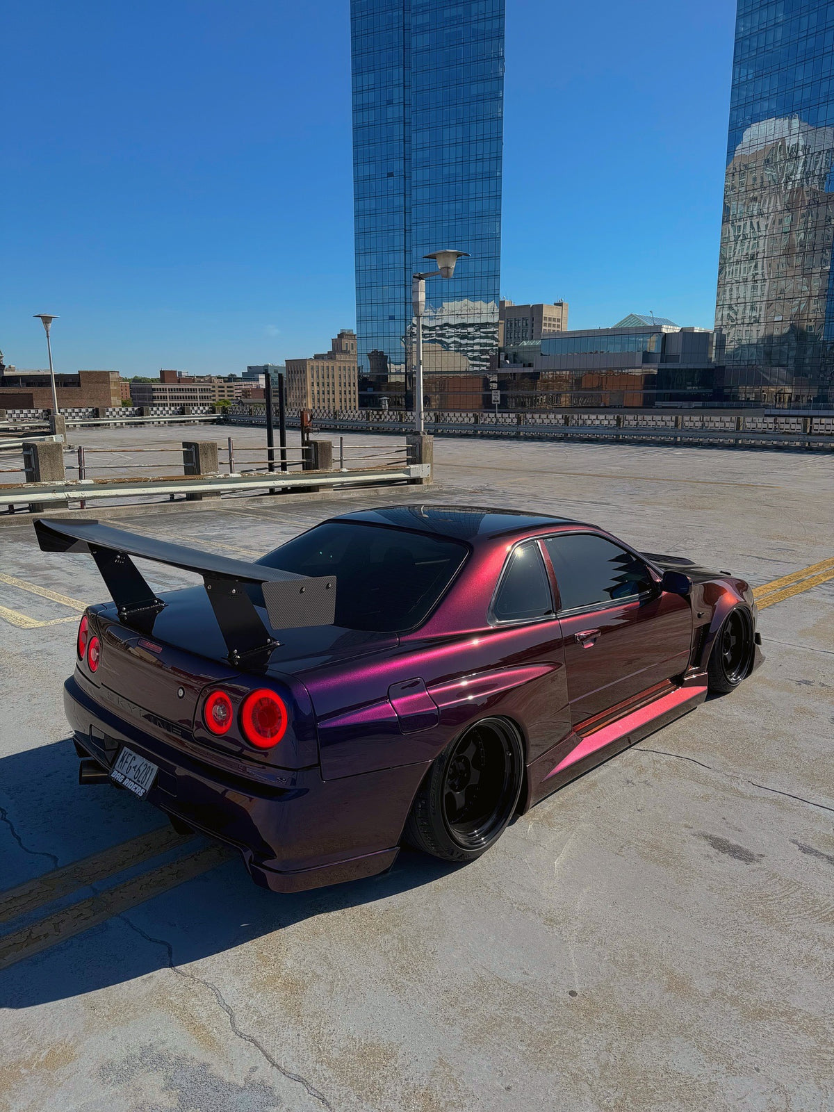 Rear three-quarter view of a color-shift purple Nissan Skyline R34 with an Epsilon+ GT wing on bottom-mount uprights, on a rooftop