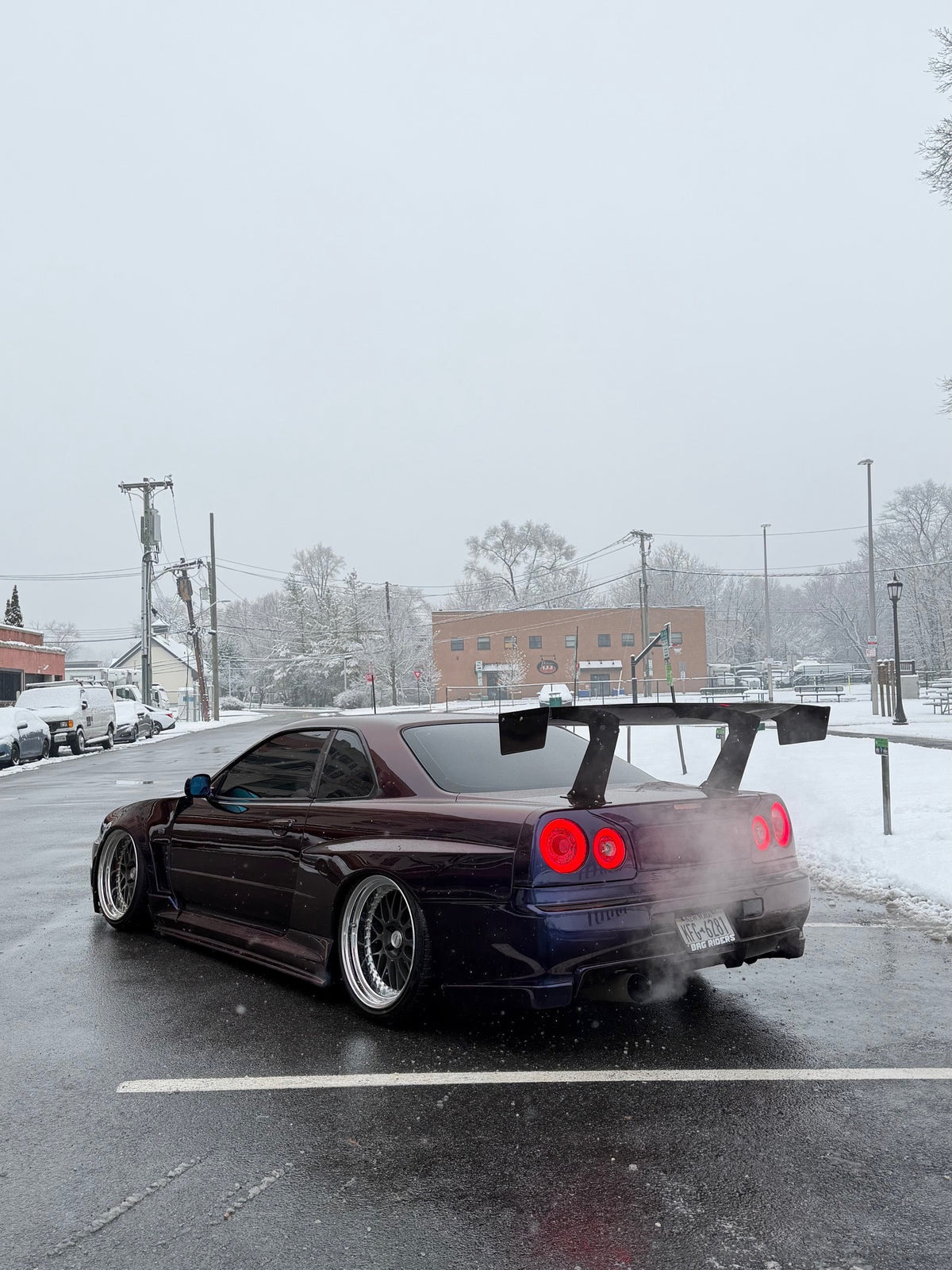 Rear three-quarter view of a purple Nissan Skyline R34 with an Epsilon+ GT wing in falling snow, exhaust steam behind