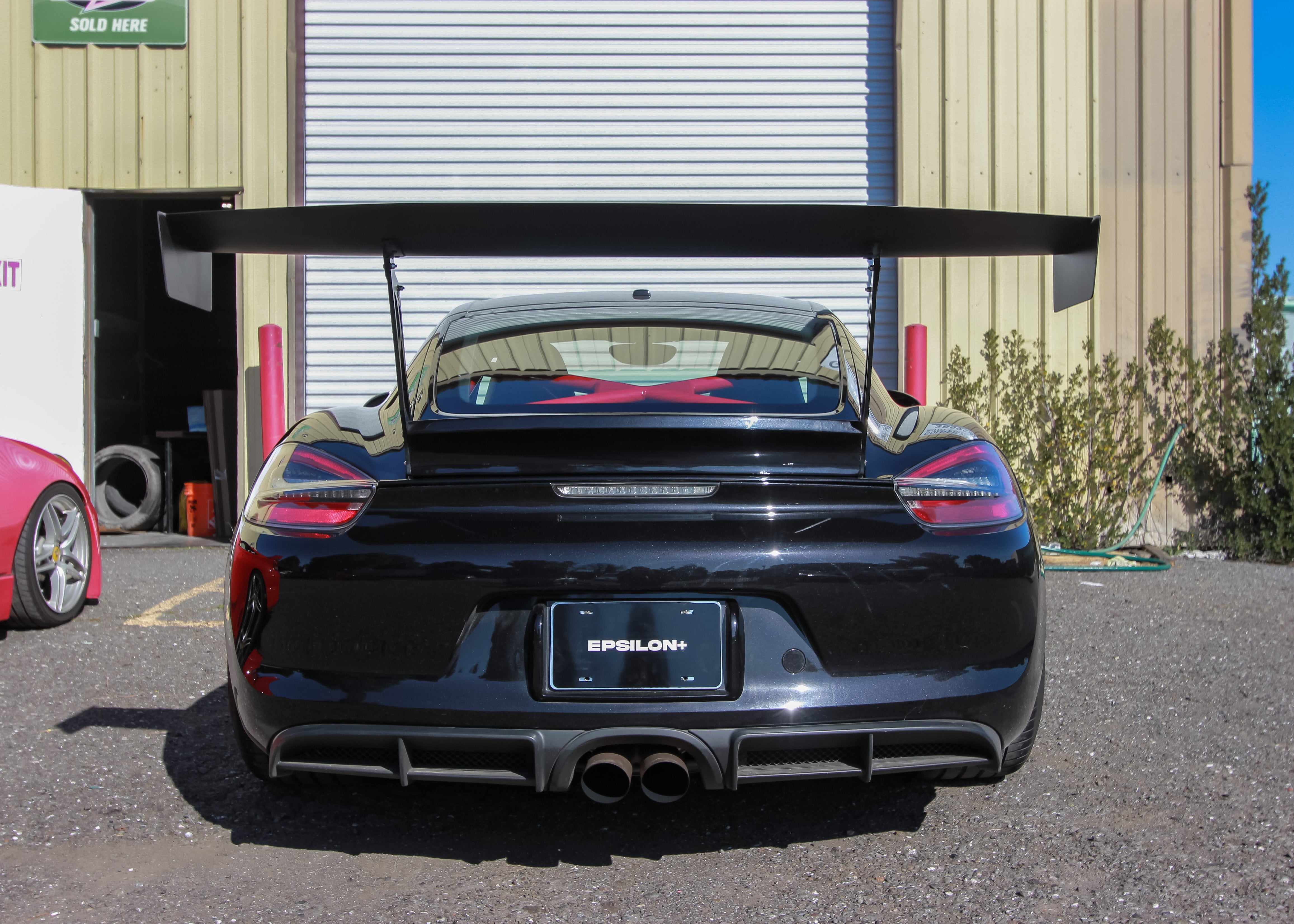 Rear view of a black Porsche Cayman showing the GT wing and Epsilon+ endplate branding