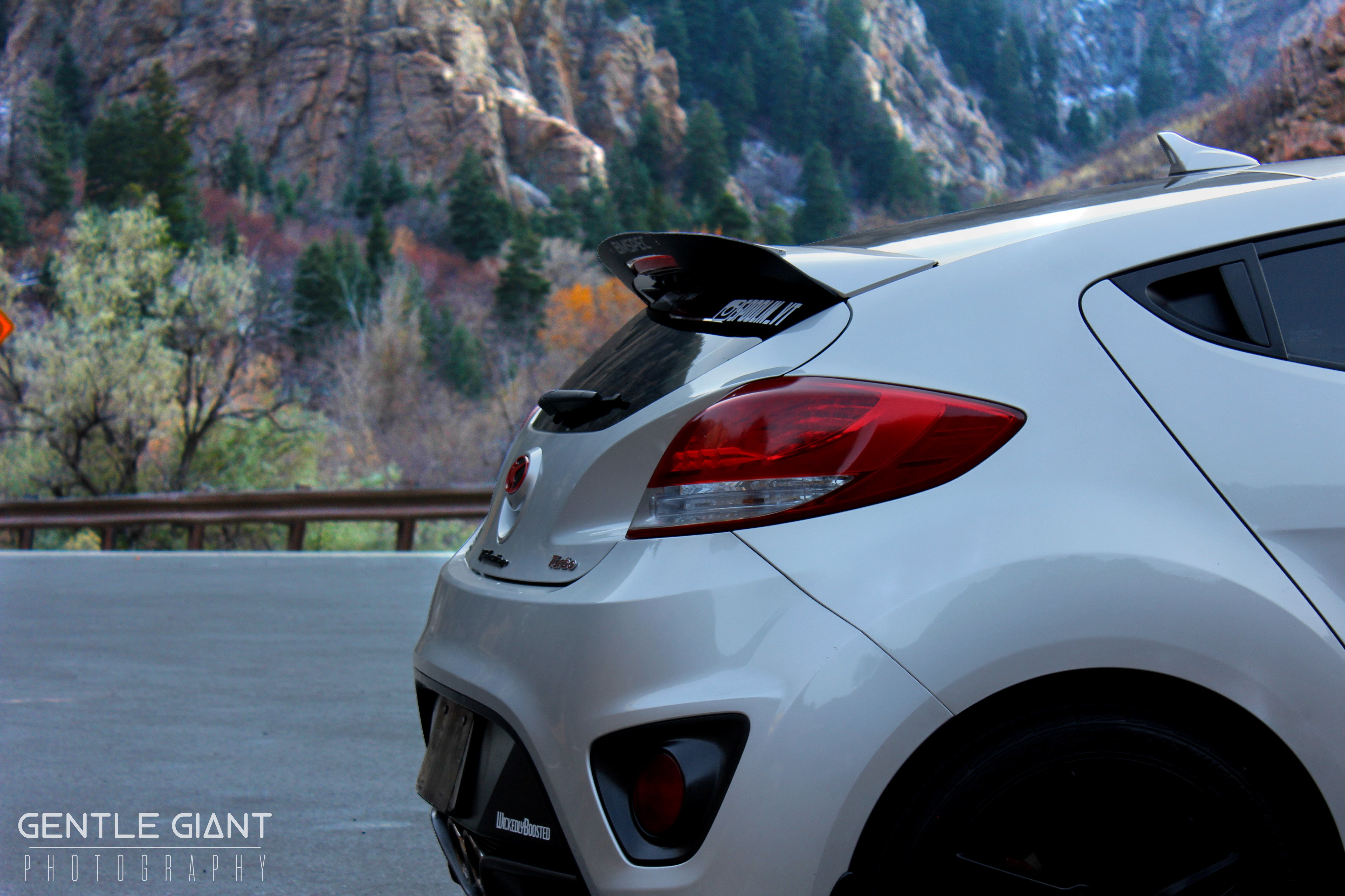 Side rear view of a white Veloster Turbo showing spoiler extension profile on a canyon road