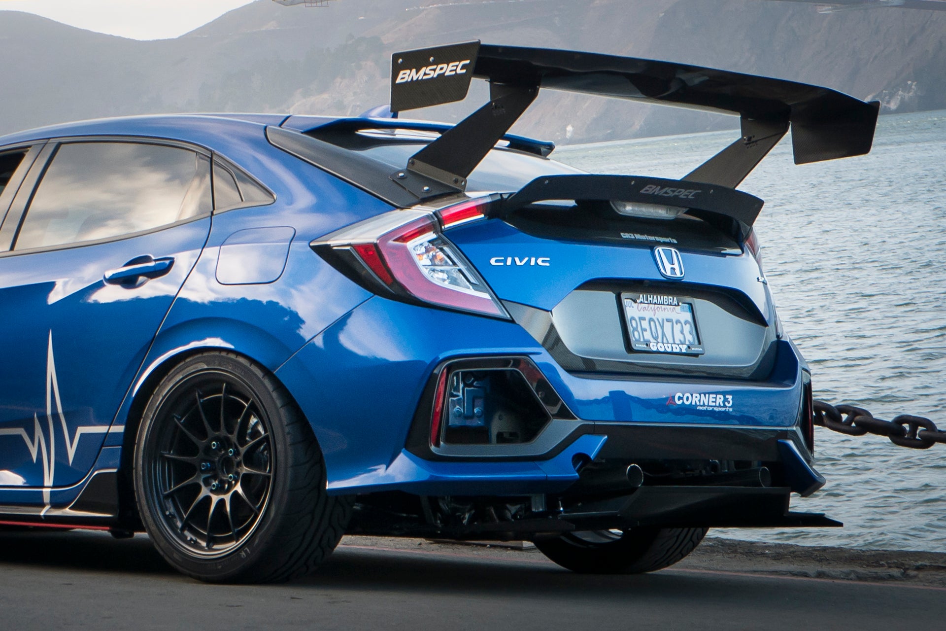 Rear three-quarter view of a blue Civic Hatchback FK7 with V1C+ diffuser beside the water
